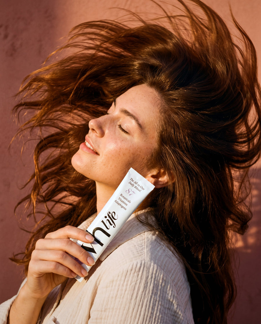 A woman with long chestnut hair lit by low sun against a terracotta wall, eyes closed, holding a VH Life sachet at her collarbone